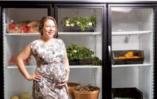 Photo of Elysia standing in front of a cooler with vegetables in it. Coolers containg fresh produce are one method of improving food access in local communities.