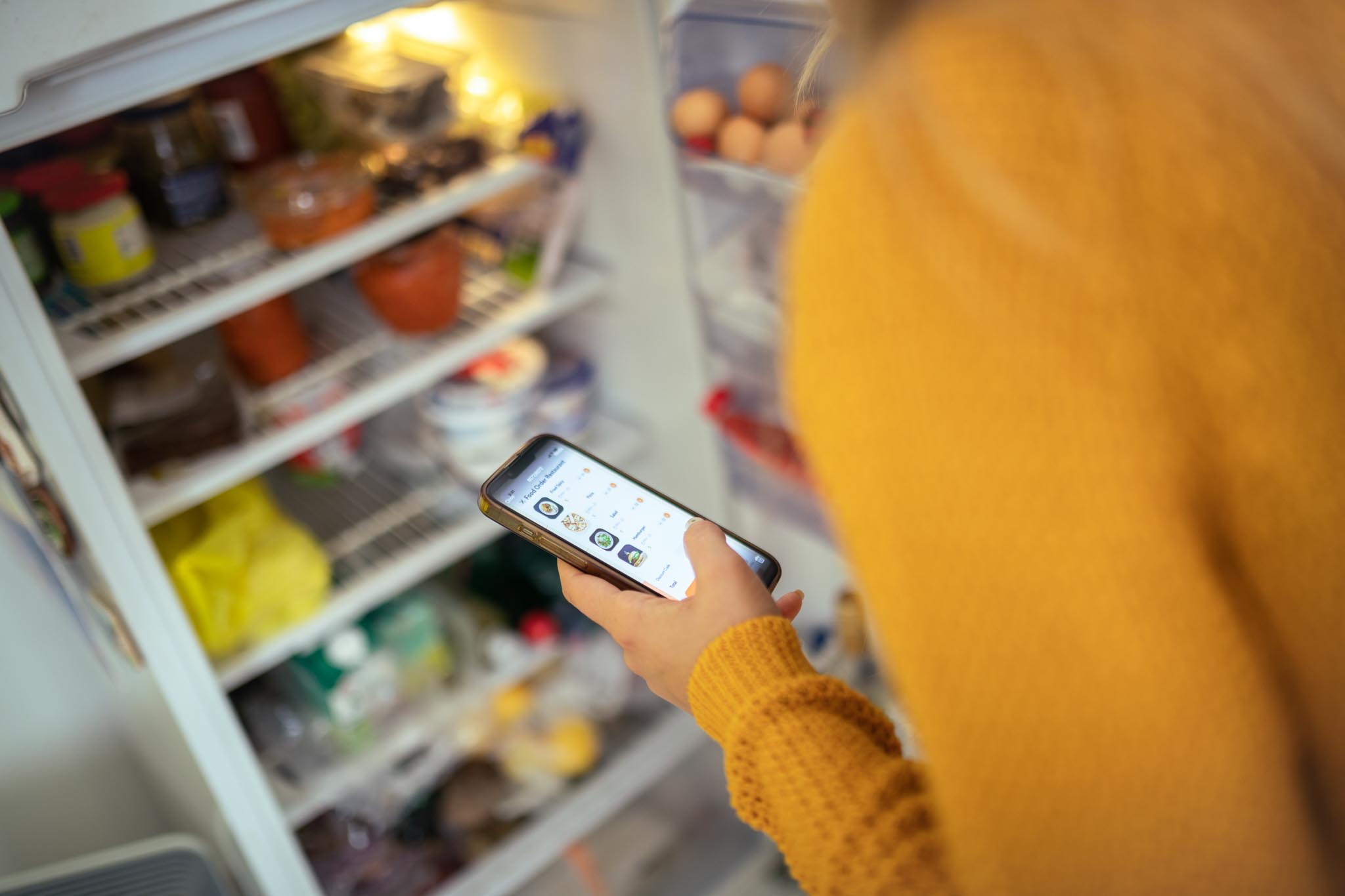 Photo of an unidentifiable person looking in a refrigerator while placing an order on their phone. Ordering online with SNAP benefits is now possible through many online retailers.